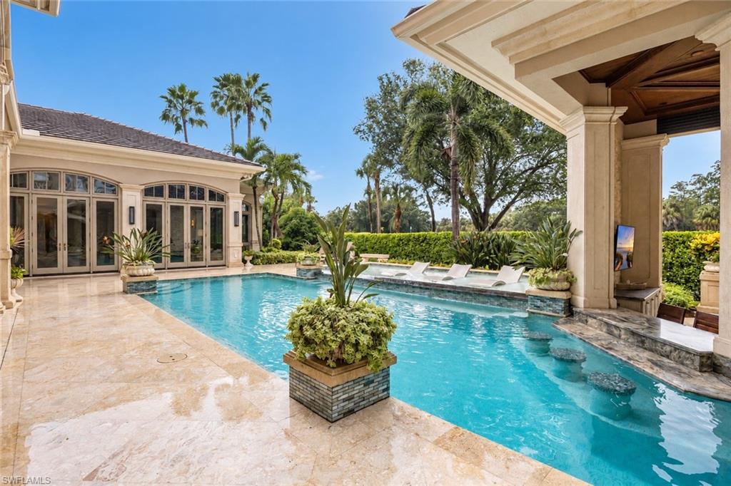 Grand architecture, a superior interior remodel and exceptional outdoor entertaining distinguish this five-bedroom plus den offering in the Estates at Bay Colony Golf Club. Featuring approximately 8,355 square feet under air, the residence welcomes guests with a sweeping staircase, soaring ceilings and beautifully appointed gathering spaces. A fireplace anchors the formal living room, where expansive windows frame the pool setting. A separate dining room, spacious kitchen, morning room, family room and wet bar accommodate both intimate evenings and larger celebrations. Dedicated media, game and wine rooms add to the home’s extensive entertaining options.

The main-level primary suite occupies its own wing with a separate retreat, multiple walk-in closets and an expansive bath. A study and second bedroom complete the main-level accommodations, while three upstairs bedrooms, a loft and balconies provide privacy for guests. An elevator connects the two levels.
Outdoor living unfolds across more than 2,000 square feet of covered lanais surrounding the pool, spa, sun shelf and sun deck. The outdoor kitchen features a wood-fired/gas pizza oven, wood-pellet smoker, Fire Magic gas grill, warming drawer and power burner, complemented by two outdoor-rated Sub-Zero refrigerators. The pool house offers a swim-up bar, gas fireplace, television, sink, two additional outdoor-rated Sub-Zero refrigerators and a cube ice maker. An outdoor-rated U-Line nugget ice maker adds convenience, while hurricane-rated shutters allow the pool house to close securely. Whole-house streaming audio and a recently installed outdoor sound system extend the experience throughout the property.

Significant updates include a new roof installed in September 2024, exterior painting in 2024, updated exterior lighting, a redone driveway and new pool controls in 2026. A 125-kW generator, two 1,000-gallon propane tanks, mature tropical landscaping and two separate garages with four-car capacity complete this thoughtfully equipped Naples estate.