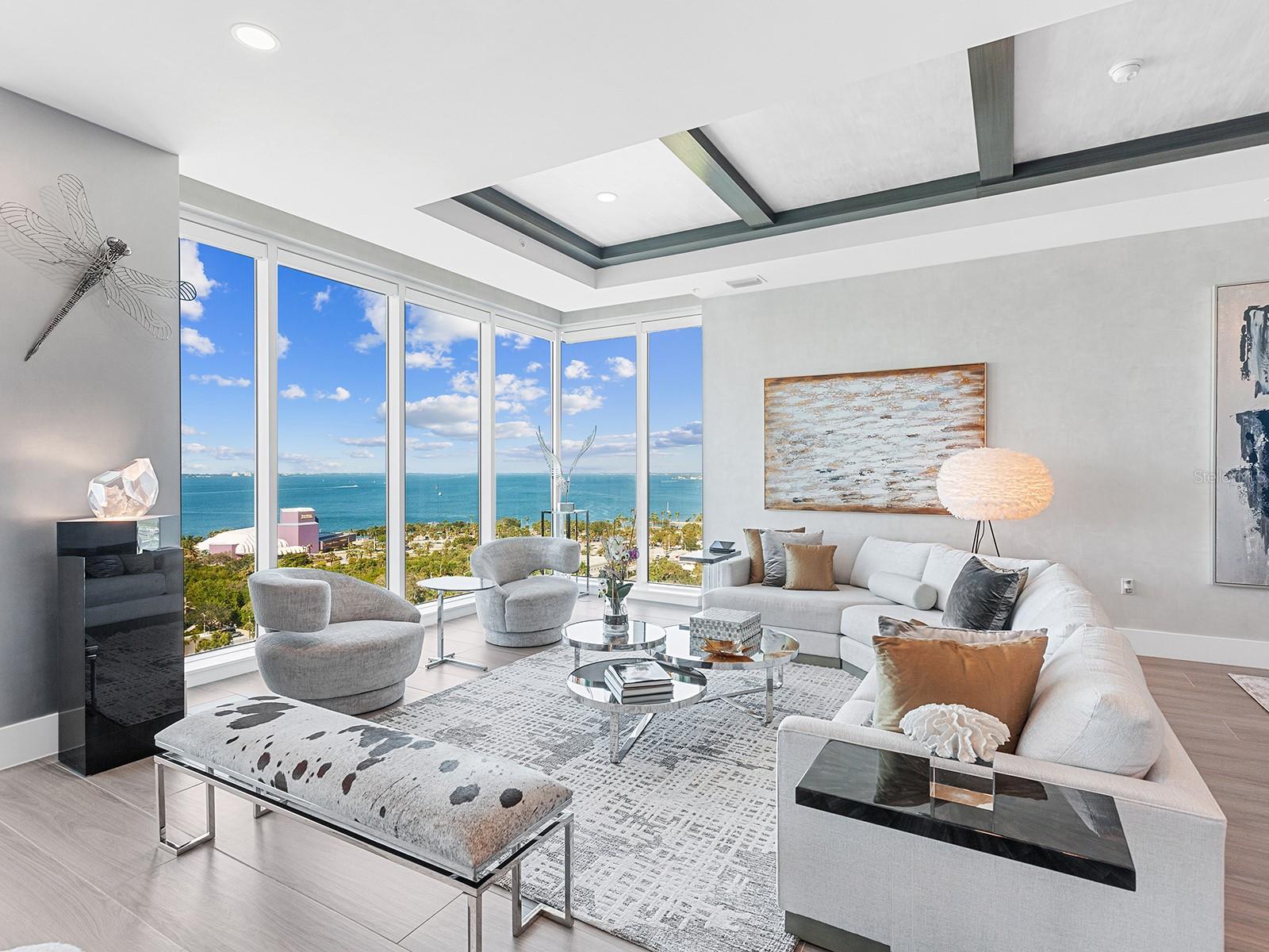 Sweeping Sarasota Bay views take center stage in this 15th-floor luxury residence at BLVD Sarasota, featured in Mansion Global. Floor-to-ceiling walls of glass wrap around the corner of the great room, revealing a breathtaking panorama of the bay, barrier islands, and open sky. As evening approaches, sunsets illuminate the water and silhouette the islands beyond, bringing extraordinary color into the living space. With three bedrooms, three private terraces, and a dedicated workspace, this downtown Sarasota condominium pairs beautifully appointed interiors with the spacious feel of a private residence and the convenience of high-rise living. A semi-private elevator opens to an elegant foyer with hand-painted walls, bespoke lighting, and wood-look plank tile flooring. The expansive great room brings living and entertaining together beneath soaring coffered ceilings, complemented by a dramatic stone accent wall with a built-in fireplace. Walls of glass connect the interior to a private balcony overlooking the city and coastline, creating an inviting setting for outdoor dining and sunset gatherings. The chef’s kitchen features custom cabinetry, quartz countertops, a designer tile backsplash, and premium Sub-Zero and Thermador appliances, including a gas cooktop and double wall oven. An oversized island incorporates seating, storage, and beverage drawers, offering generous space for meal preparation and casual dining. A built-in bar with glass shelving and a beverage cooler enhances the home’s entertaining capabilities. The primary suite offers floor-to-ceiling windows, its own private terrace, and two custom-designed walk-in closets. The spa-inspired primary bathroom includes a quartz vanity, soaking tub, glass-enclosed walk-in shower, and expansive windows that fill the space with natural light. Two additional guest suites provide privacy for visitors, while a dedicated workspace accommodates working from home. A separate laundry room with extensive cabinetry adds practical storage and everyday convenience. Three private terraces extend the living space outdoors, with sweeping views reaching toward Siesta Key, Lido Key, and Longboat Key. From morning coffee overlooking Sarasota Bay to evenings framed by the illuminated skyline, the residence offers a distinctive perspective on Florida’s Gulf Coast. BLVD Sarasota residents enjoy a rooftop deck with a zero-horizon swimming pool, a fitness center, club and wine rooms, an indoor golf simulator, and dedicated dog amenities, including a grooming salon and dog park. Located in downtown Sarasota, this luxury condominium brings together panoramic bay views, private outdoor living, designer finishes, and resort-style amenities.