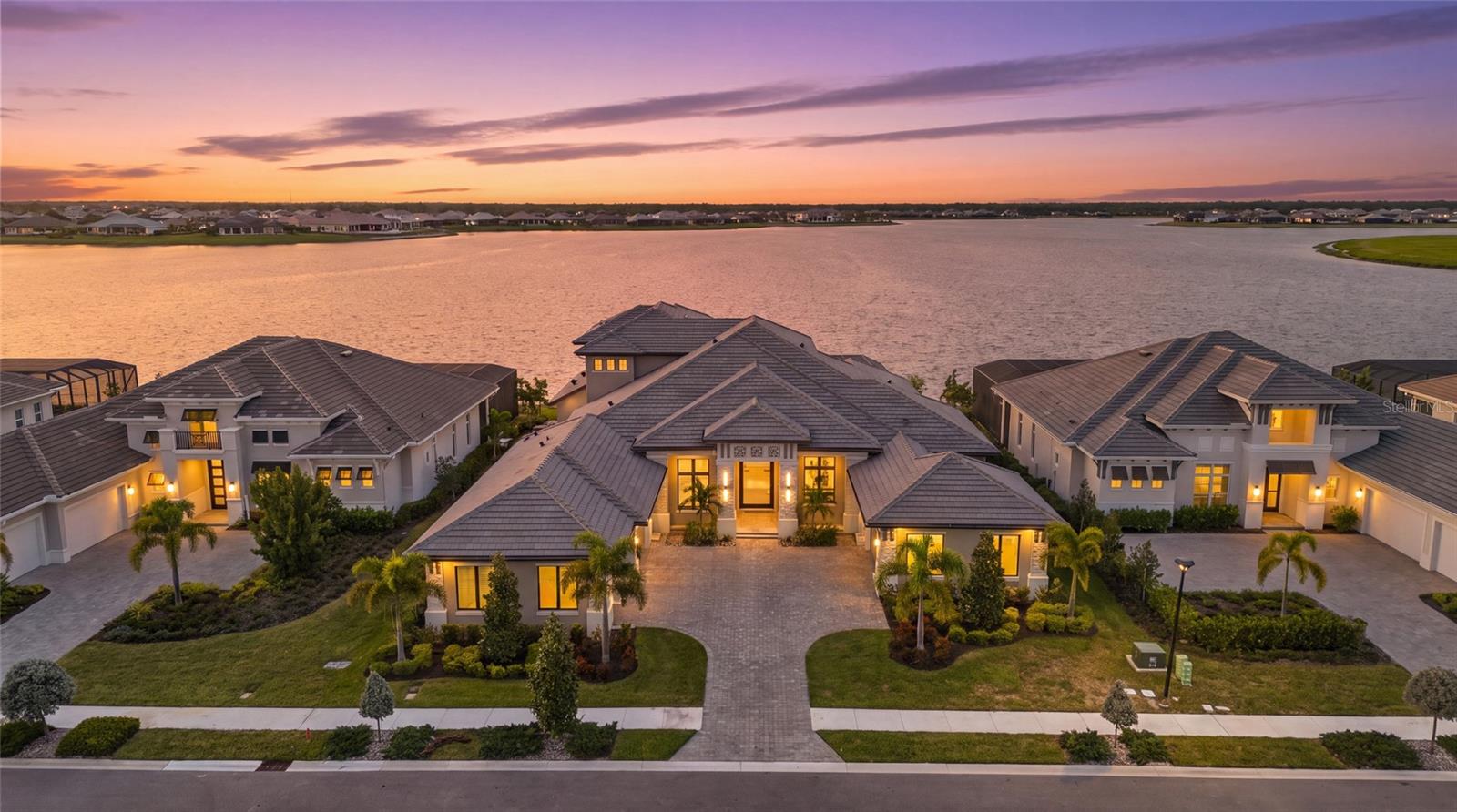 Set on one of the prime lakefront homesites within highly sought-after Wild Blue at Waterside, this completed custom John Cannon residence offers the rare opportunity to move right in and enjoy nearly 6,000 square feet of luxury living without waiting close to two years to design and build from the ground up. Complete with a five-car split garage, sweeping lake views, and exceptional indoor-outdoor living, the home stands out within one of Lakewood Ranch’s premier waterfront communities. From the oversized entry to the soaring ceilings, architectural details, wood flooring, and walls of impact glass, the home makes a striking first impression. A dedicated office near the entry provides a refined workspace. Nearby, a secluded first-floor guest or in-law suite offers an ensuite bath and direct connection to a secondary screened lanai positioned between the suite and first-floor bonus room, adding flexibility for guests and everyday living. The heart of the home centers around a dramatic great room with decorative beams and expansive sliders framing the water. A custom entertainment wall with an electric fireplace creates a striking focal point, complemented by automatic window treatments and seamless access to the outdoor living areas. Adjacent to the great room, the kitchen is designed to match the scale of the residence with Thermador appliances, custom cabinetry, an oversized center island, generous workspace, and substantial windows that bring in natural light. A large walk-in pantry provides exceptional storage while allowing the primary kitchen to remain polished and streamlined for entertaining. Additional bedrooms feature ensuite accommodations, while a dedicated laundry room adds everyday functionality without compromising the home’s clean architectural flow. The primary retreat is positioned to maximize the waterfront setting, with oversized windows showcasing the lake and coffered ceiling details overhead. The suite extends into generous dual bath and dressing spaces with oversized showers and expansive walk-in closets, creating a true owner’s retreat with privacy and scale. Upstairs, a second bonus room creates a separate living and entertainment space. An ensuite bedroom with a walk-in closet is ideal for guests or extended stays, while the expansive screened upper balcony captures elevated panoramic lake views. Back downstairs, impact sliders open to a substantial lanai with a full outdoor kitchen overlooking the lake. The resort-style pool and spa incorporate fire and water features, dramatic stepping stones, and an ultra-view screened enclosure designed to preserve the view. A gas connection is already in place for a future fire pit. For the automotive enthusiast or collector, the five-car split garage offers substantial space with elevated ceiling heights and potential for vehicle lifts. Beyond the residence, Wild Blue at Waterside offers a premier resort-style lifestyle with recreation, fitness, sports, trails, social amenities, and convenient access to Waterside Place for shopping, dining, and entertainment. With the home completed and ready to enjoy, 596 Blue Shell Loop offers the opportunity to step directly into the Wild Blue lifestyle without waiting through the custom-build process. A rare blend of custom craftsmanship, commanding lake views, resort-style outdoor living, and five-car garage capacity, this is a residence that leaves a lasting impression.