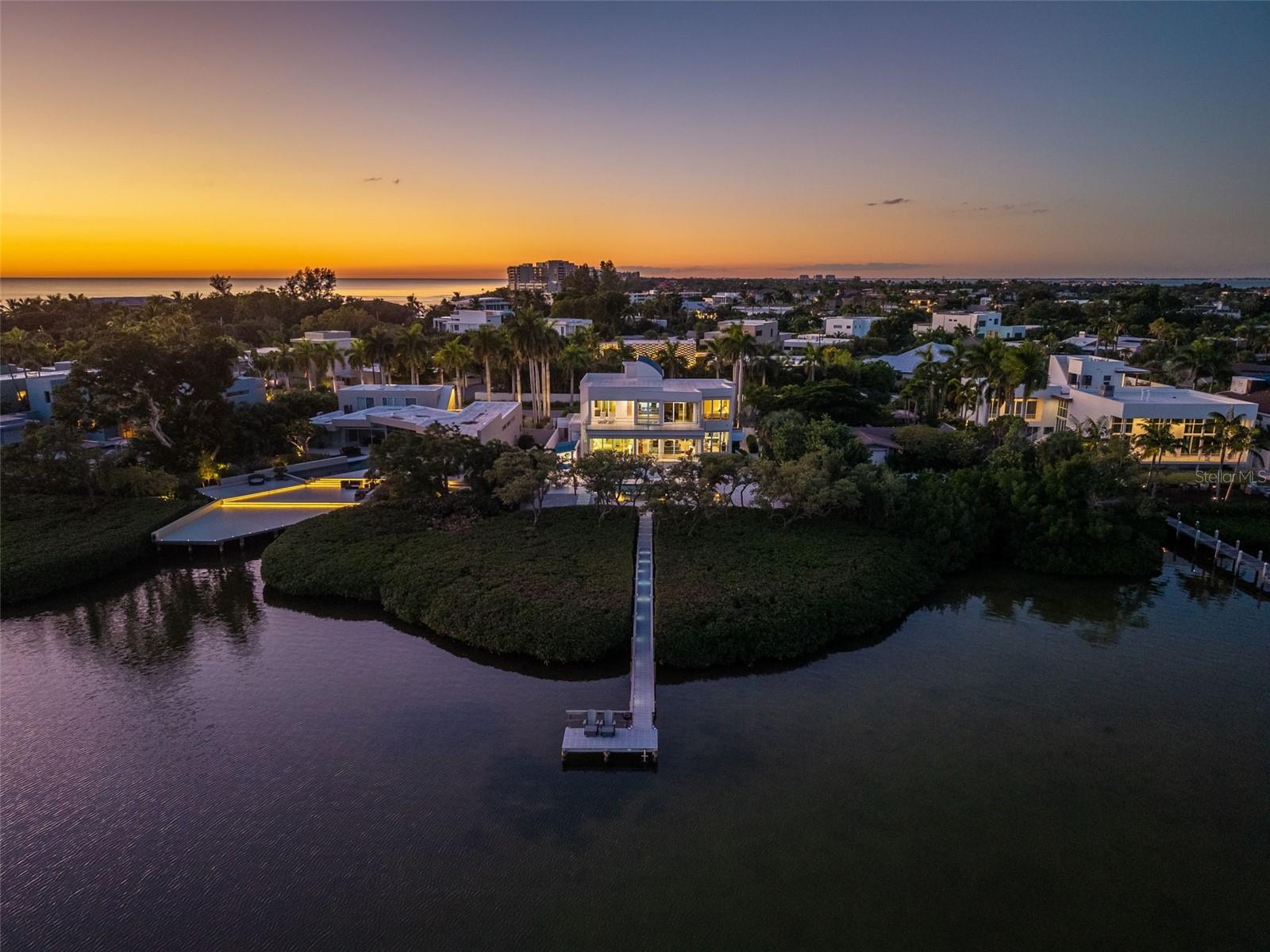 Mornings begin with light moving across the bayou. Coffee on the terrace, a swim in the infinity-edge pool, and time to enjoy the quiet before heading to St. Armands Circle or the beach. Set along the waterfront in Lido Shores, this custom-designed modern residence pairs bold architectural lines, expansive glass, and warm natural materials with the tranquility of its bayou setting. From the circular brick-paver drive, cedar accents, architectural trellises, and tropical plantings offer a welcoming introduction. Inside, the home opens to a great room with nearly 20' ceilings, where walls of glass draw your attention straight to the water. The scale is striking, yet Beechwood floors and natural cedar bring warmth and texture to the expansive spaces. Throughout the day, changing light and reflections from the bayou become part of the interior. The kitchen provides a natural gathering place, with an illuminated onyx island at its center. It is easy to picture friends collecting here over a glass of wine while dinner takes shape. Porcelanosa cabinetry and quartz countertops create a clean architectural backdrop, complemented by a paneled Sub-Zero refrigerator, Wolf wall oven, warming drawer and microwave, and a walk-in pantry.
With 4,284 square feet of living space, five bedrooms, and three-and-a-half baths, the home offers room to come together and places to retreat. Two primary suites, one on each level, enjoy water views, private terraces, spacious walk-in closets, and beautifully appointed baths, including one with a steam shower. From the upstairs terrace, views extend across the water to the Ringling Bridge and downtown Sarasota skyline, offering a beautiful backdrop as the city lights come on each evening. Comfortable guest accommodations welcome family and friends, while a dedicated office and waterfront fitness room make even the quieter routines of the day feel connected to the setting.
Outside, the terraces unfold toward an infinity-edge saltwater pool that appears to meet the bayou beyond. An integrated spa and cascading planter waterfall add movement and sound, creating a setting that invites you to stay a little longer. Lunch beneath the covered lanai can stretch into an afternoon by the pool, followed by dinner outside as the light softens over the water. Electric screens offer added comfort, and a separate pavilion with gas and water connections is ready for a future summer kitchen. An illuminated dock provides a departure point for exploring Sarasota’s waterways, with access to Sarasota Bay. Set on approximately .37 acres, the residence also features impact-rated windows and doors, automatic solar window treatments, imported European lighting, integrated LED illumination, smart-home technology, and a dedicated laundry room. Home to some of the Sarasota School of Architecture’s most prized modern homes, Lido Shores has a character shaped by its architectural heritage and the people who live here. Neighborhood gatherings, potluck dinners, and familiar faces create a sense of belonging, where getting to know your neighbors is part of the appeal. Private deeded access to Lido Key Beach offers a quiet place to gather at the end of the day and take in some of Sarasota’s finest sunsets. With St. Armands Circle, Longboat Key, and downtown Sarasota just minutes away, you can enjoy everything the area offers while coming home to a neighborhood that feels distinctly its own.
