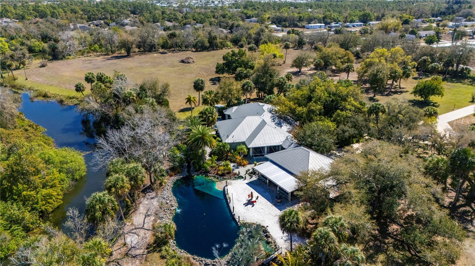 Privacy, waterfront living, acreage & exceptional flexibility come together on 3.7 acres along serene Alligator Creek in Punta Gorda. Beautifully designed private estate home offers more than 3,500sqft of living space, generous outdoor living areas, substantial garage & storage space & a peaceful setting surrounded by Florida-friendly landscaping. It also offers additional possibilities, including the potential to function as an Airbnb, to be a wedding & memorial venue for up to 35 events annually, & offers real estate density for up to 17 units. For those seeking an even larger opportunity, two adjacent 5 acre parcels are available separately, or the entire 13 acres w/ the home may be purchased together, offering 67 units of density for a single-family or manufactured home community. Beyond the gated entrance, this owner-designed, structurally reinforced concrete-built estate unfolds. A spacious front porch leads to the double-door entry, where crown molding, tray ceilings, transom windows & generous living areas create an impressive sense of space. The flexible floor plan includes formal living & dining areas, an executive office, family room/game room, & a large kitchen featuring top-of-the-line appliances, a center island, abundant solid wood cabinetry, breakfast bar, and granite countertops. Some furniture is included, while additional furniture & game room equipment are negotiable. An oversized laundry room provides additional storage, while the split-bedroom floor plan creates privacy between the primary suite & guest accommodations. The private primary suite features split vanities, a jetted soaking tub, separate walk-in shower, & large his-and-hers walk-in closets. Three guest rooms, three full baths, an outdoor half bath, & a separate heated outdoor shower complete the accommodations. Two screened rear patios extend the living space outdoors and provide peaceful places to take in the quiet surroundings. From there, the grounds become an experience of their own. Tennessee flagstone walkways wind through the property to an approximately 80,000-gallon contained natural outdoor lagoon/pond with a concrete bottom and walk-in sandy beach area. Custom rock fountains, waterfalls, & a koi pond add layers of water features throughout the backyard, creating a distinctive setting for relaxation, entertaining, and enjoying the Florida sunshine. Car enthusiasts, collectors, or those needing substantial workshop & storage space will appreciate the detached 38x30-foot air-conditioned 4+ car garage w/ three electric overhead doors. The property also includes an additional 30x15-foot storage room w/ two overhead doors, a 30x15 covered patio & entertainment area, & a Hot Springs hot tub. The waterfront setting along Alligator Creek adds another dimension to this Southwest Florida estate, w/a private dock & access to the natural beauty and outdoor lifestyle Punta Gorda is known for. Despite its peaceful and secluded atmosphere, yet convenient to shopping, dining, I-75 and Tamiami Trail, with easy access to fishing, boating, Charlotte Harbor, golf, pickleball, tennis, trails, entertainment and historic downtown Punta Gorda. Award-winning beaches -Boca Grande, Englewood Beach and Manasota Key are also within easy reach. Whether enjoyed purely as a private Punta Gorda waterfront estate or explored for its Airbnb, venue, acreage, and development potential, this property offers a rare combination of lifestyle, privacy, flexibility, and opportunity.